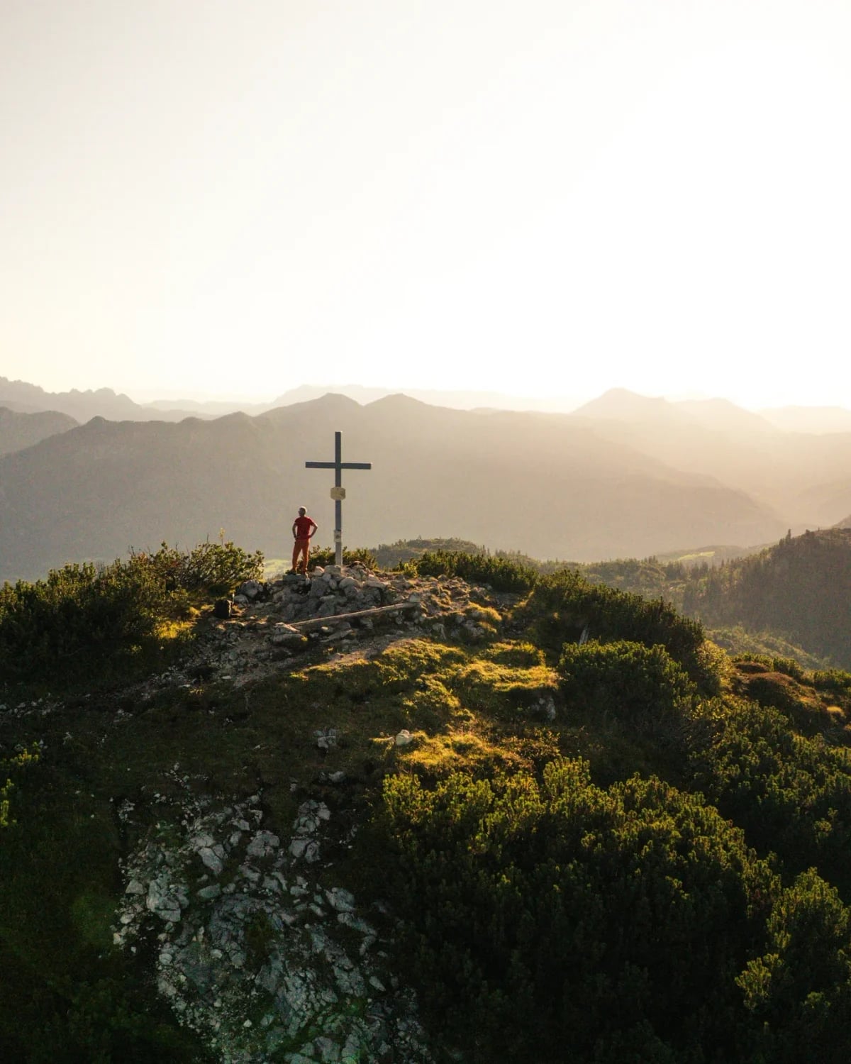 Drohnenaufnahme: Person am Gipfelkreuz im Gegenlicht, dahinter Bergketten im Dunst der Abendsonne.